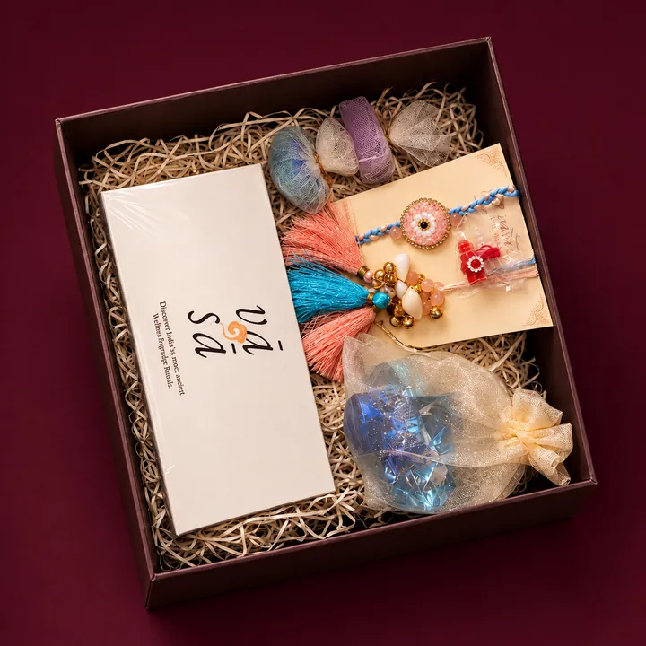 An open VASA box packed in straw with rakhi threads on a card, a crystal in a sheer pouch and a white VASA box